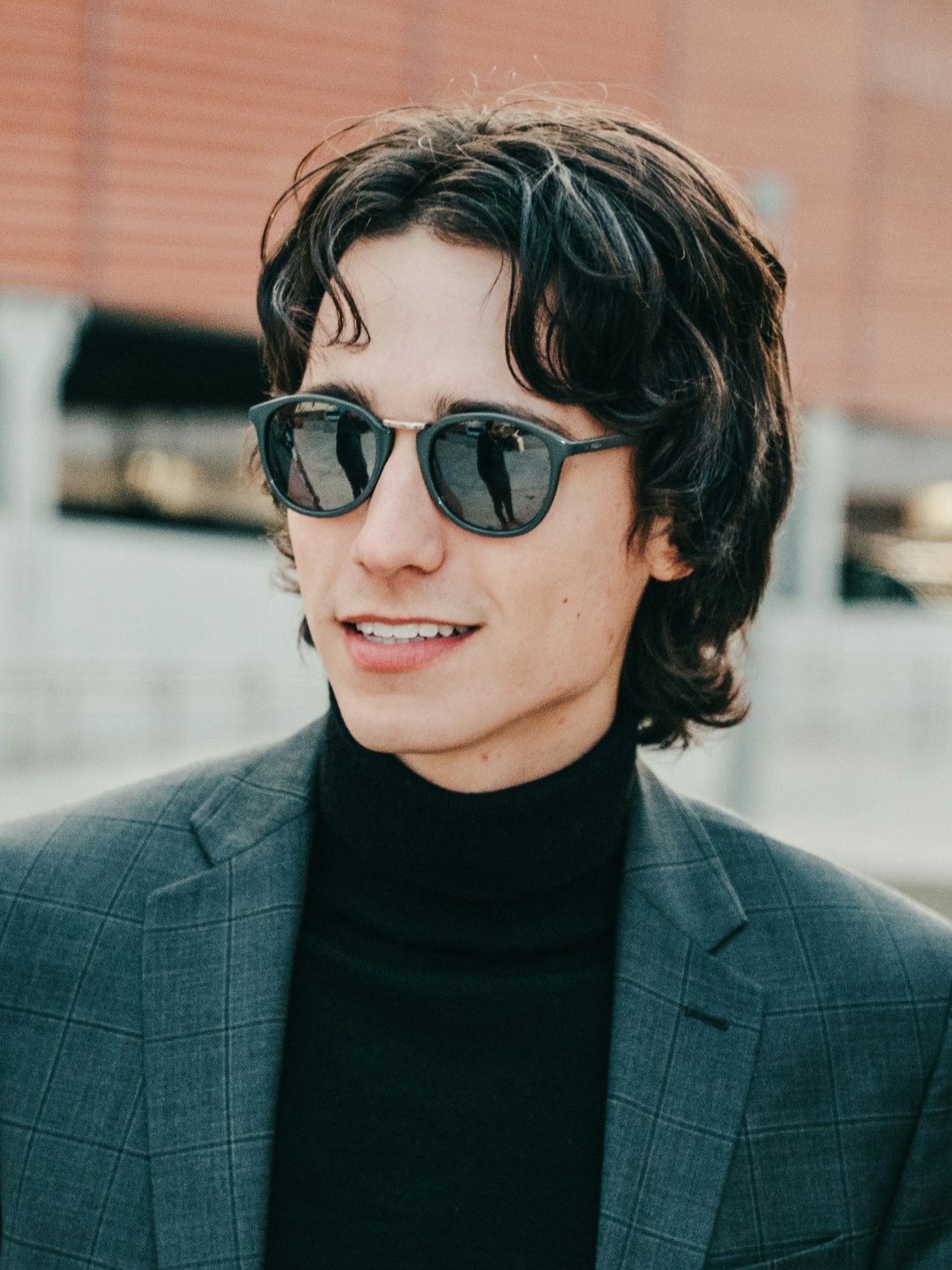 Un jeune homme avec une coupe mulet rétro, portant une veste de costume grise et des lunettes de soleil, souriant en extérieur.