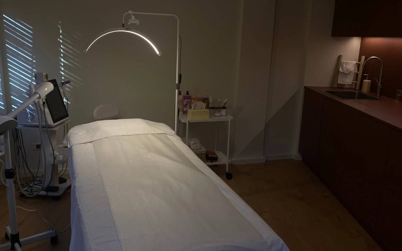 Une salle de soin focalisée avec un lit blanc sous une lampe à arc LED, un appareil d’esthétique professionnelle avec écran numérique, des produits de soin sur un chariot, et des meubles en bois sombre avec un lavabo le long du mur du fond.