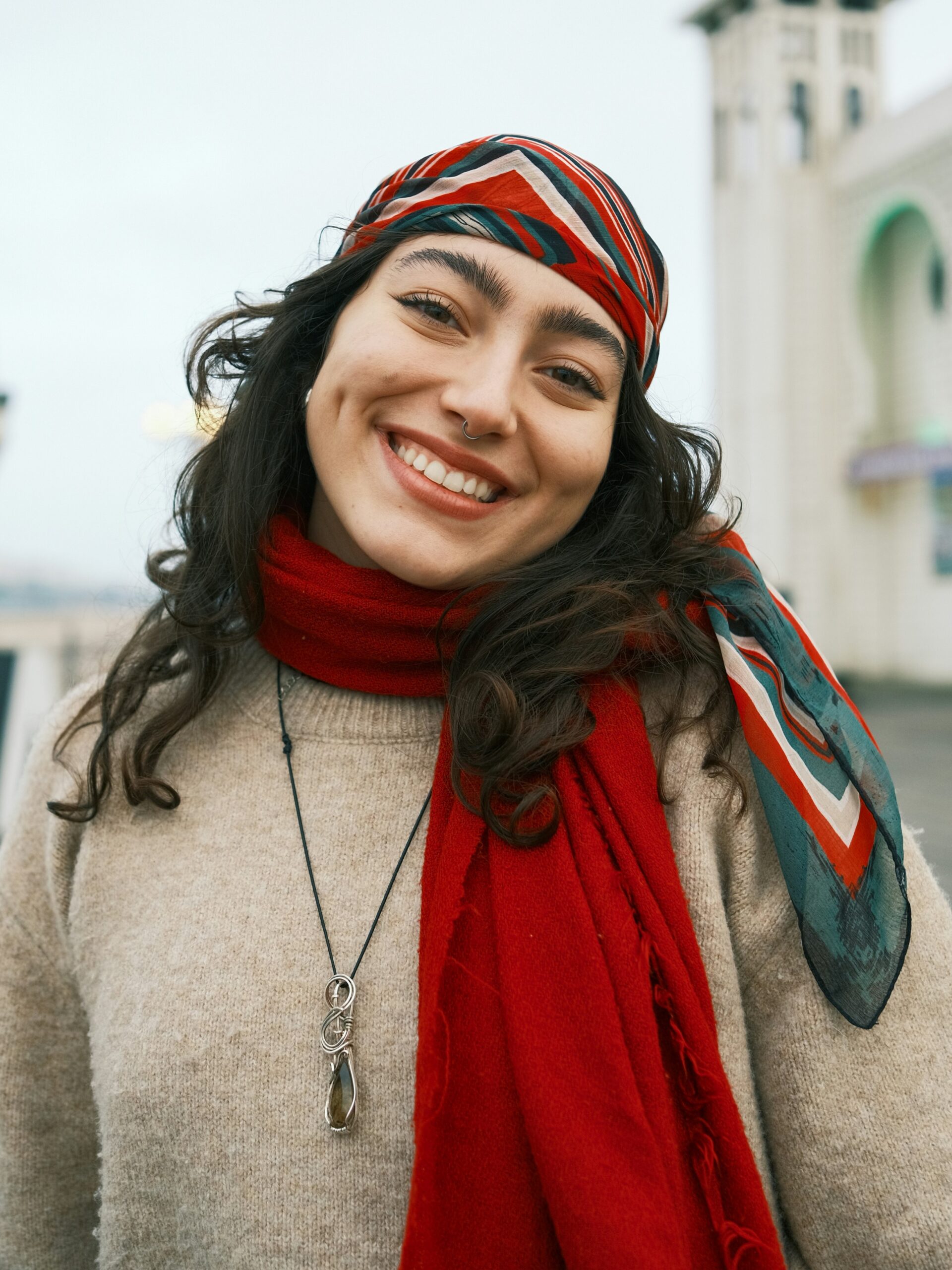Femme souriante debout sur une jetée, avec de longs cheveux bruns, portant un foulard rayé rouge, blanc et vert sur la tête et une écharpe rouge autour du cou