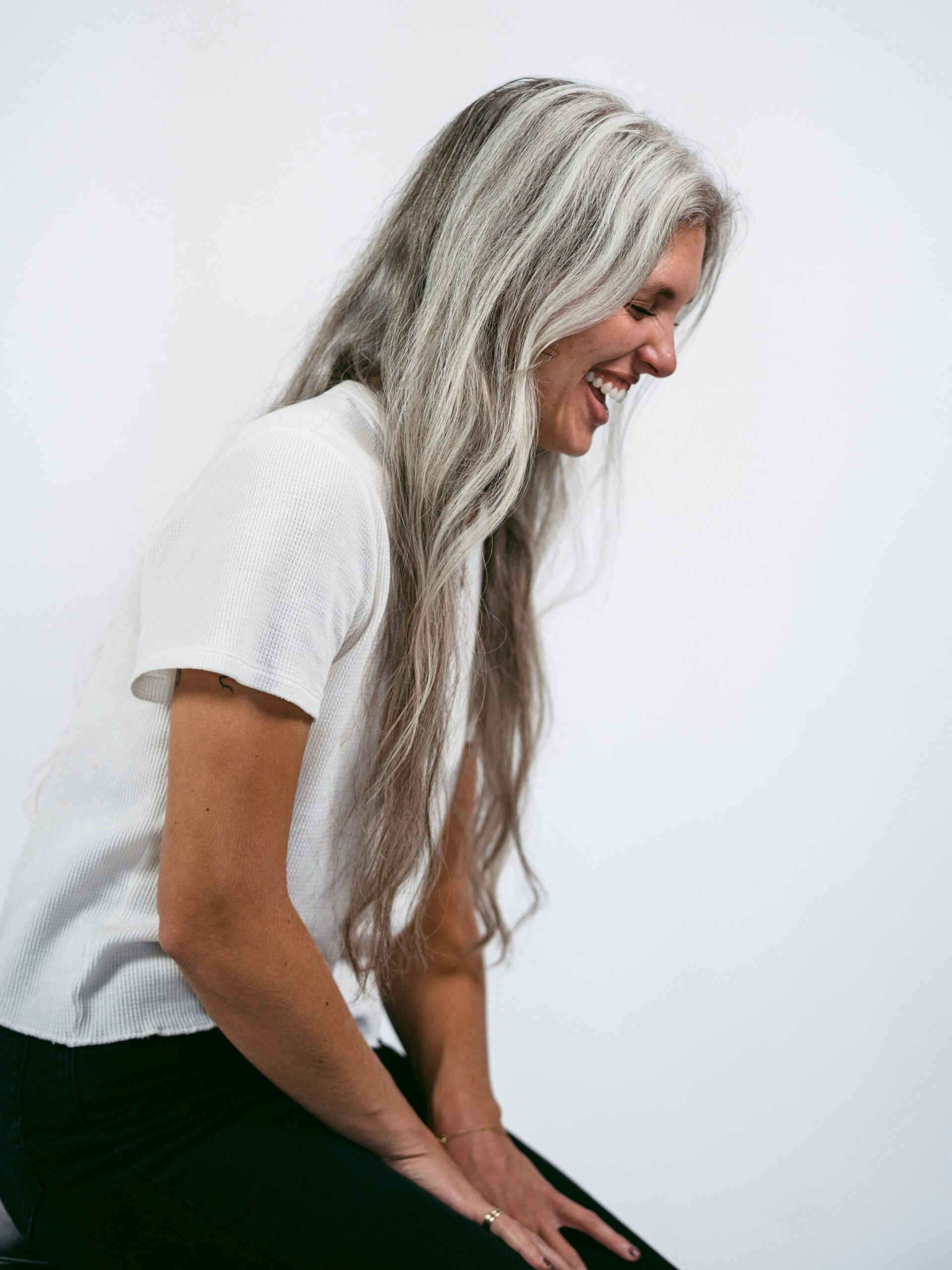 Vue de profil d’une femme assise sur un tabouret, riant, avec de très longs cheveux gris
