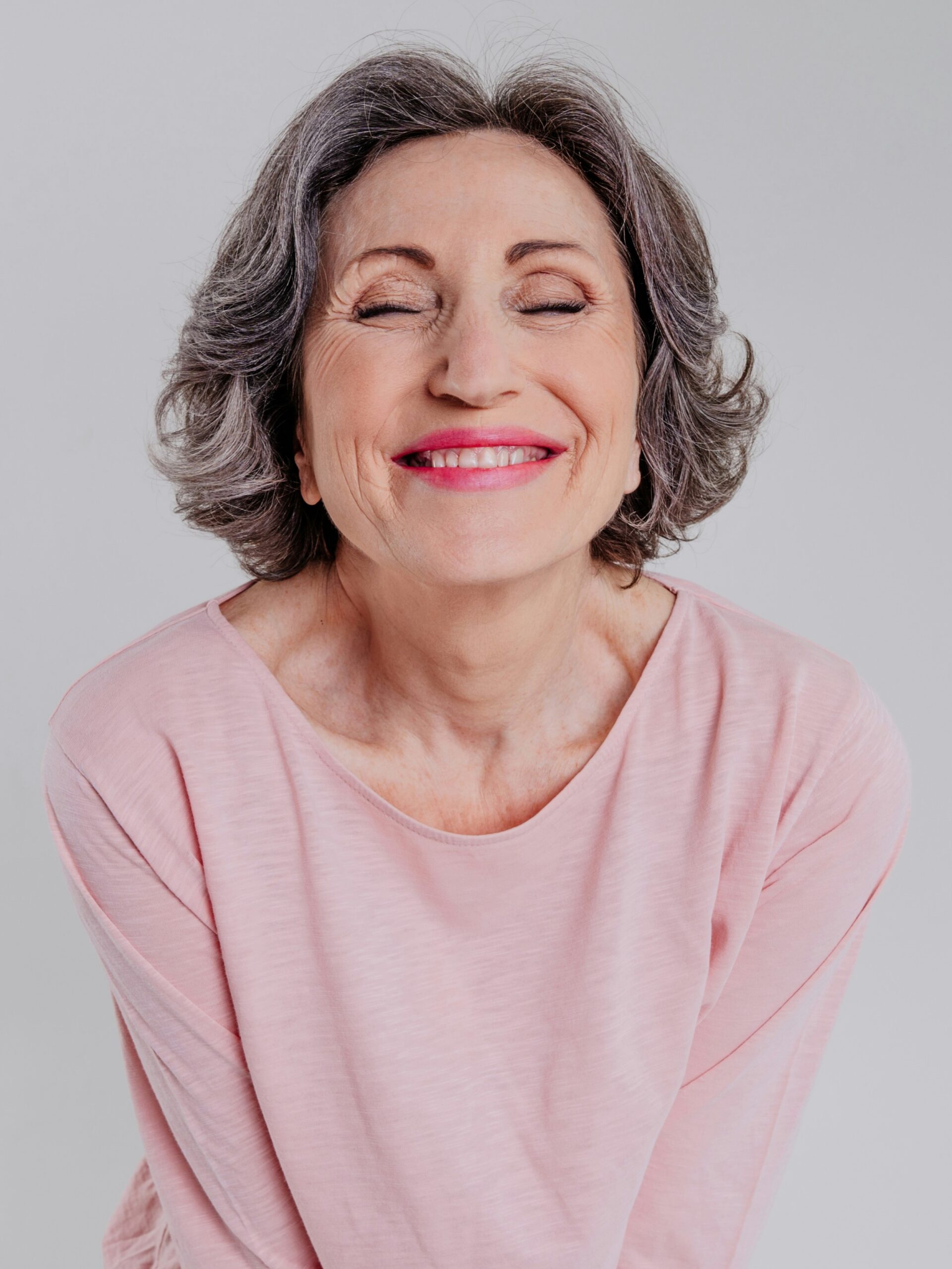 Portrait d’une femme aux cheveux courts et ondulés poivre et sel, portant un haut rose ample et souriant les yeux fermés