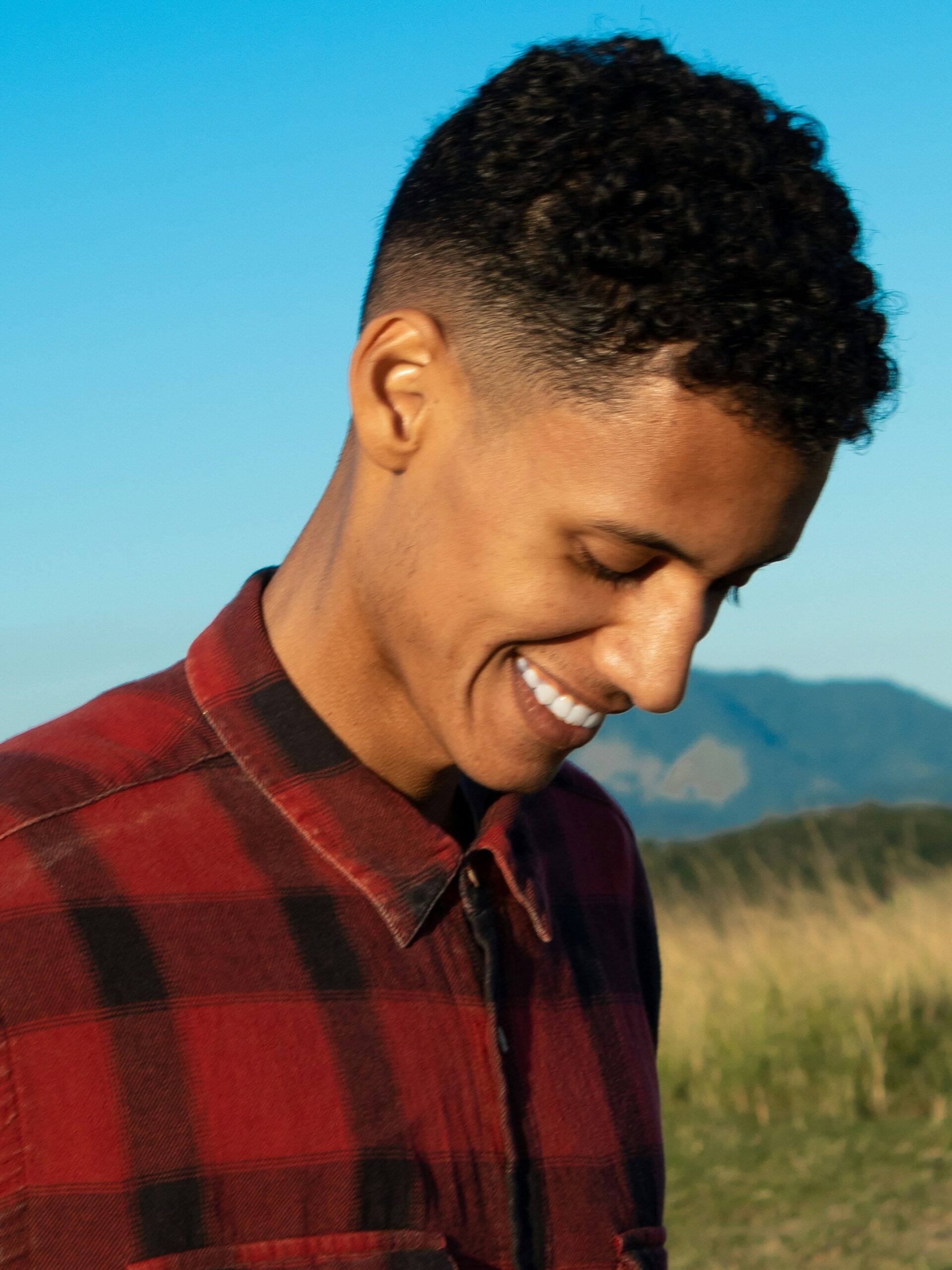 Homme souriant en extérieur devant un paysage de montagnes et de ciel bleu, avec des cheveux bouclés et un taper fade moyen