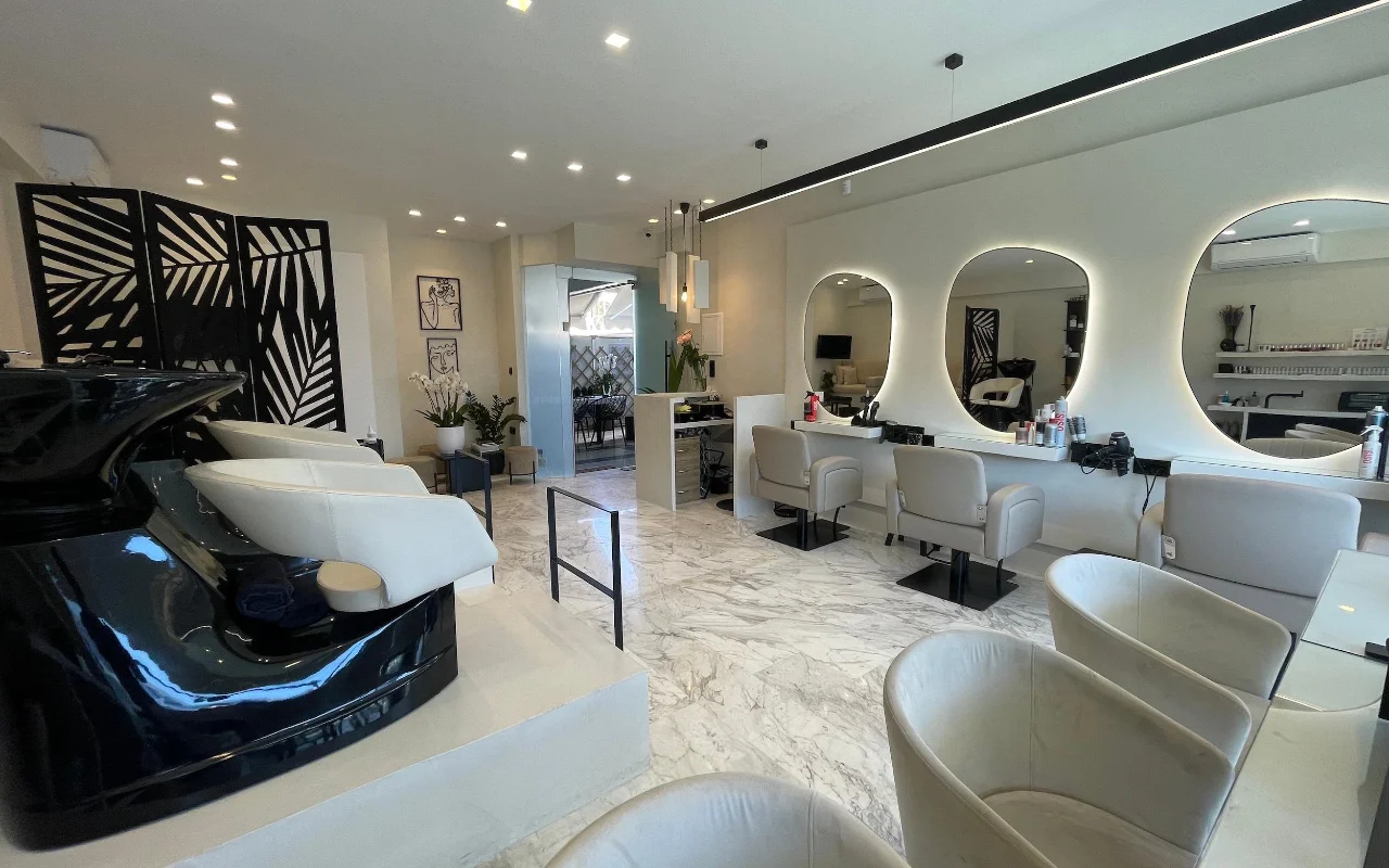 Un salon de coiffure élégant avec des sols en marbre blanc, des fauteuils de coiffage en cuir crème sous des miroirs ovales cerclés de LED, un bac à shampoing noir laqué, et un paravent géométrique à motifs de feuilles de palmier.