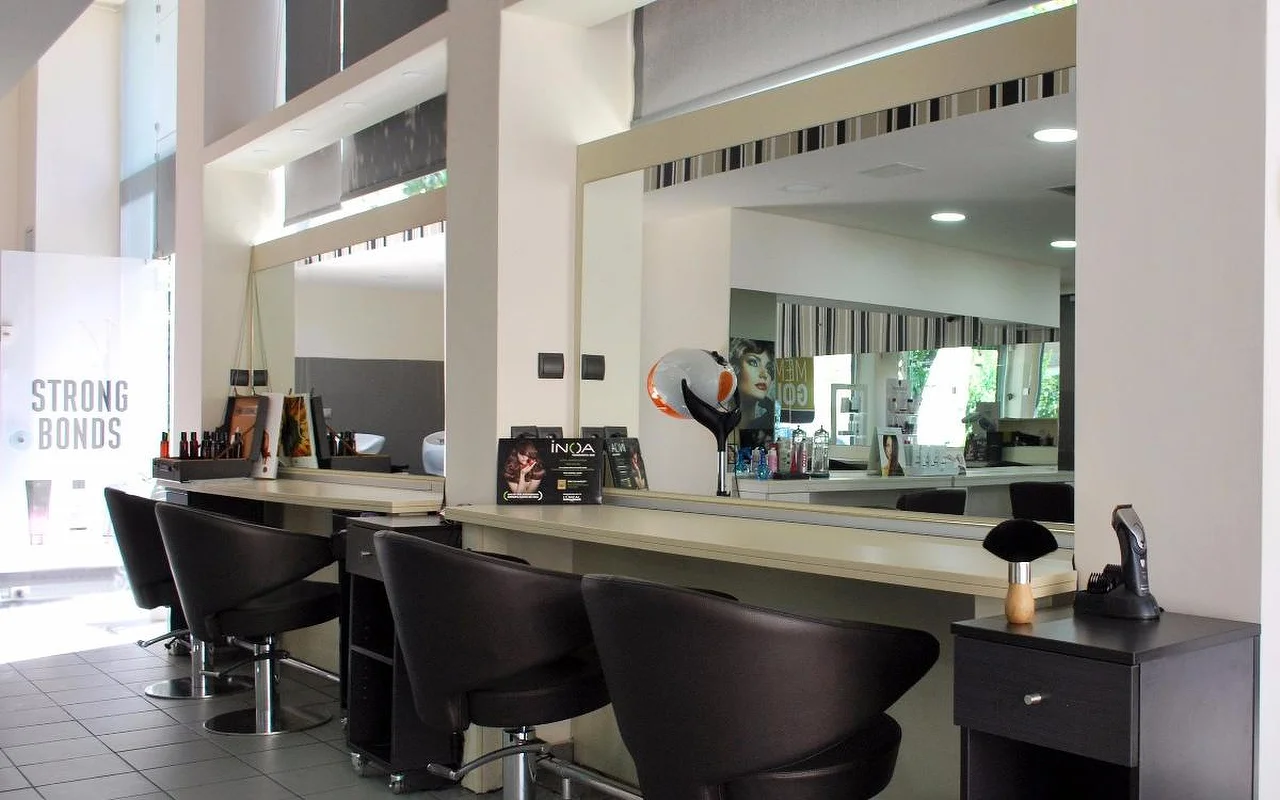 Un salon de coiffure professionnel avec des fauteuils de coiffage en cuir sombre face à de grands miroirs, des produits capillaires alignés le long du comptoir, de hauts plafonds et une lumière naturelle filtrant par des impostes vitrées.