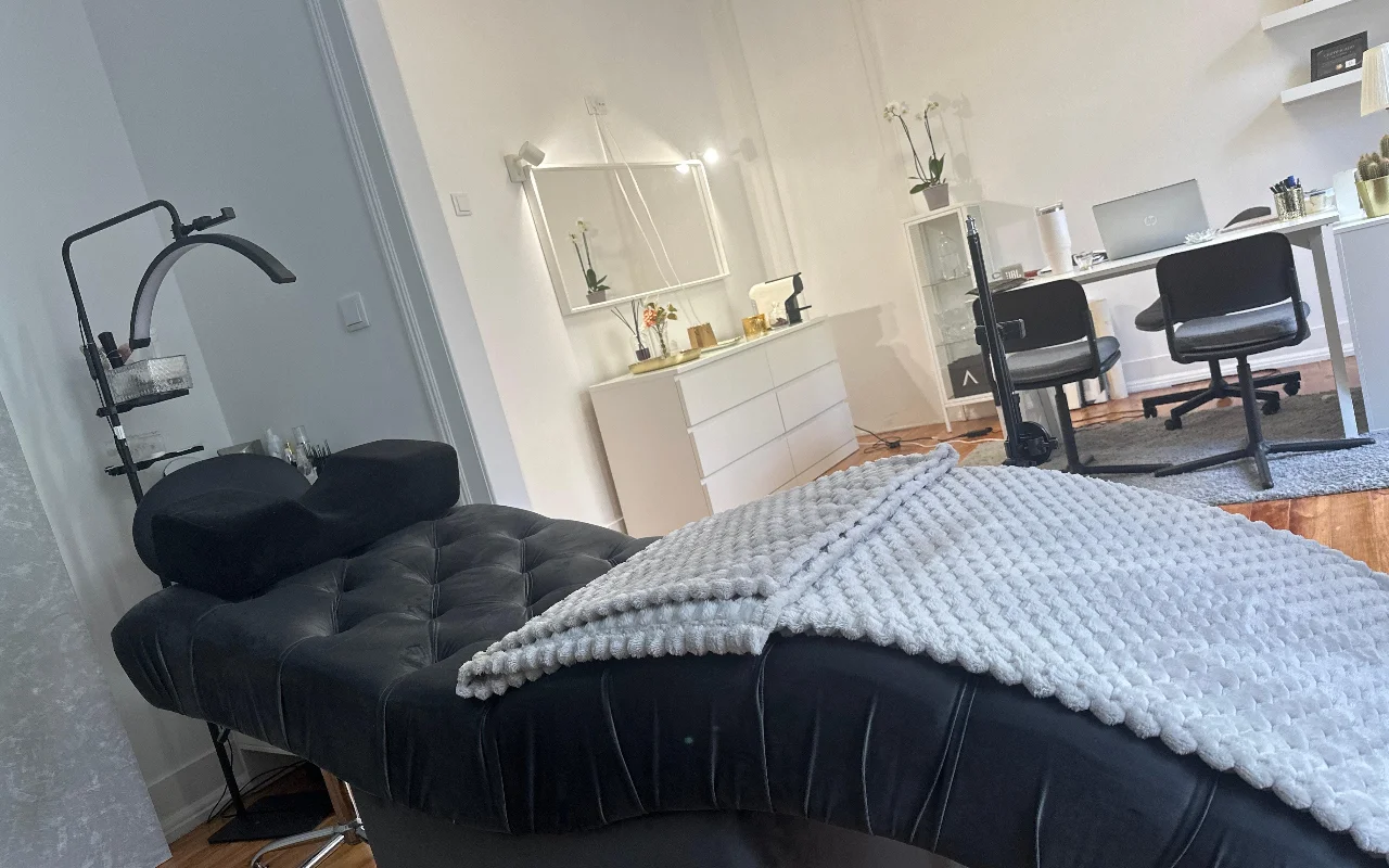 Une cabine de soin des cils moderne photographiée en contre-plongée. Au premier plan, une méridienne capitonnée noire est drapée d'un plaid gris texturé et équipée d'un coussin ergonomique sous une lampe annulaire courbée. En arrière-plan, un bureau minimaliste blanc avec un ordinateur portable, deux chaises noires, une commode blanche et un grand miroir.