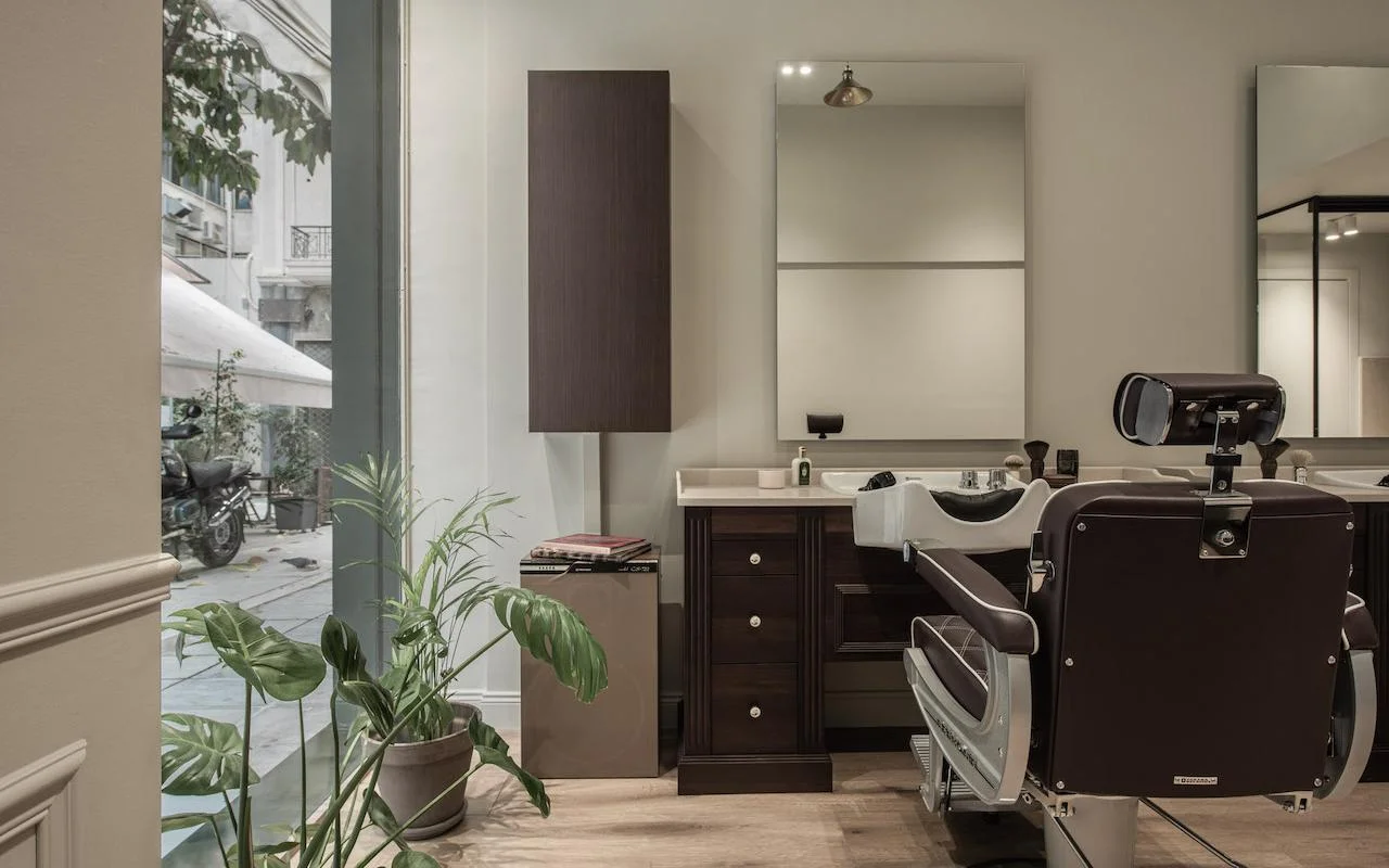 Un barbershop raffiné avec un fauteuil de barbier en cuir brun foncé, des meubles en noyer, une grande fenêtre donnant sur une rue de Kolonáki, et un monstera apportant une touche de verdure dans l’espace minimaliste.