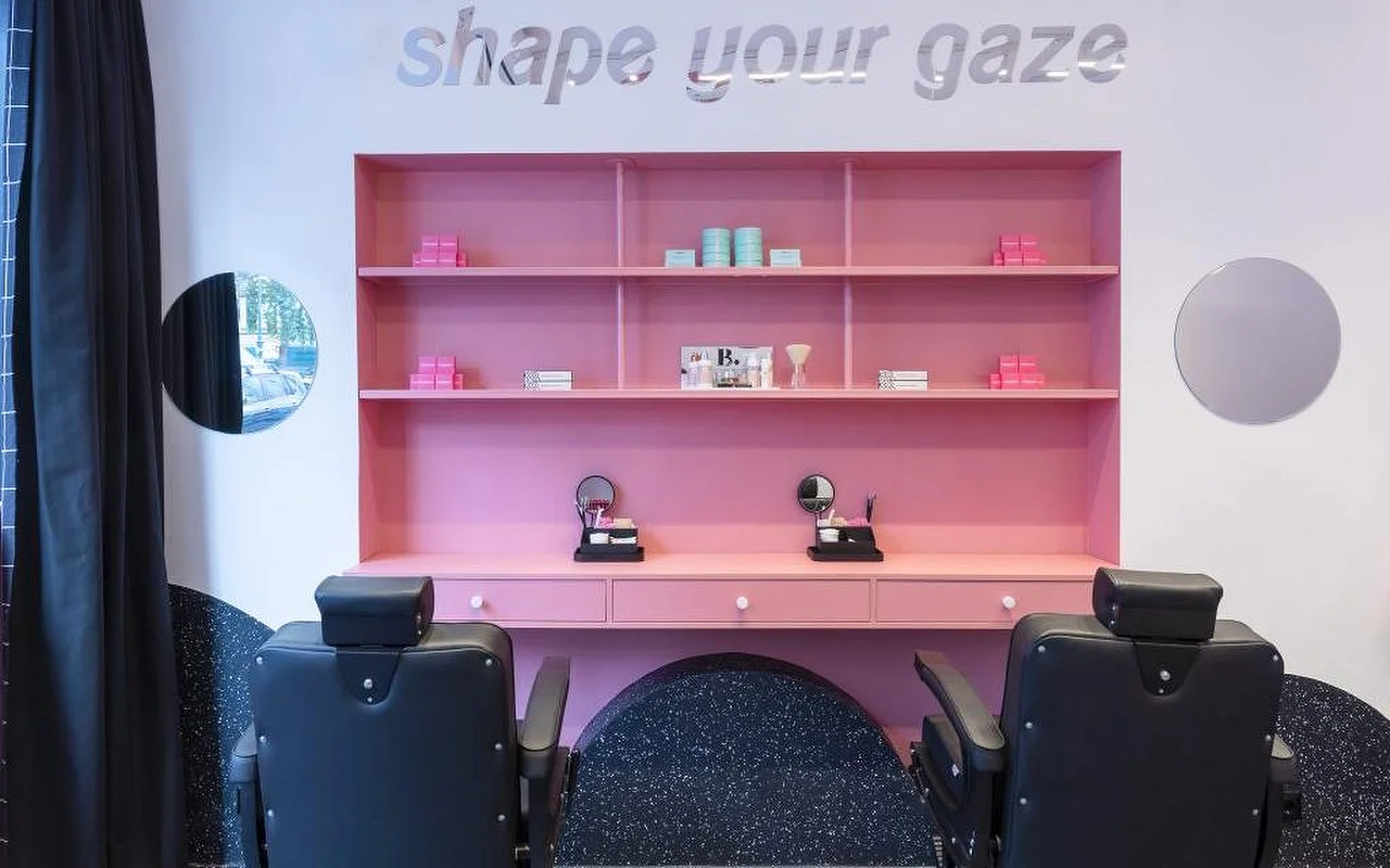 Un studio spécialisé sourcils et cils avec une étagère rose vif présentant des produits de beauté, un lettrage argenté « shape your gaze » au mur, et des fauteuils de soin en cuir noir sur un sol moucheté sombre.