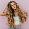 Photo studio d'une femme aux très longs cheveux ondulés effet soleil réalisés en balayage, vêtue d'un jean bleu clair et d'un top court en maille blanc