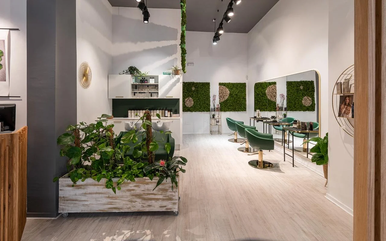 Un salon de coiffure rempli de plantes aux sols en bois clair, un grand pot blanc patiné débordant de verdure, une œuvre murale en mousse végétale, des fauteuils en velours vert émeraude aux détails dorés, et un bac de lavage à l'arrière.