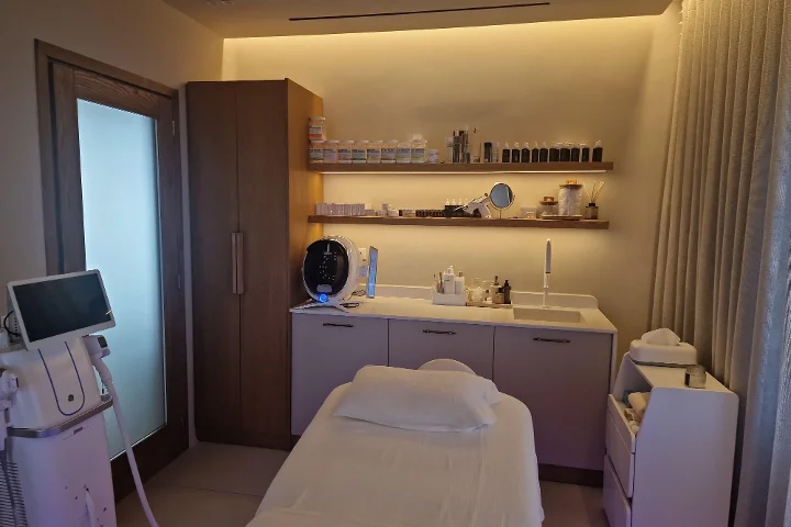 Une cabine de soin à la lumière chaude, avec une table de soin du visage blanche et des serviettes pliées avec soin, une étagère en bois foncé garnie de produits de soin, un écran d'analyse de peau et des appareils de traitement à côté, clinique mais cosy.
