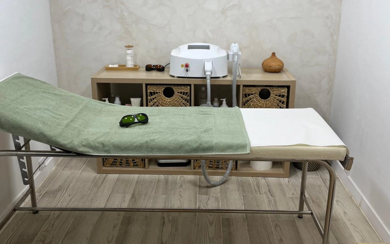 Une cabine de soin aux tons neutres avec une table à structure métallique recouverte d'une serviette vert sauge, une paire de lunettes de protection foncées posées dessus, un appareil laser sur une console en bois, et des paniers de rangement tressés en dessous.
