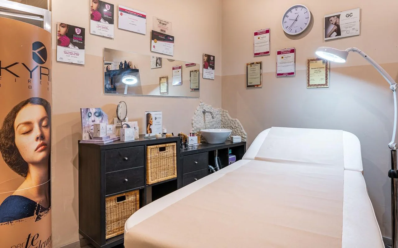 Une cabine de soin chez Oltre la bellezza, avec une table de soin blanche, une lampe-loupe réglable et des murs tapissés de certificats et de diplômes professionnels. Des meubles de rangement en osier foncé sont disposés sous un miroir, et des produits de soin sont alignés sur le plan de travail.