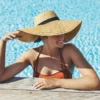 Une femme dans une piscine extérieure ensoleillée, accoudée au rebord, portant un bikini orangé et un chapeau de paille à large bord