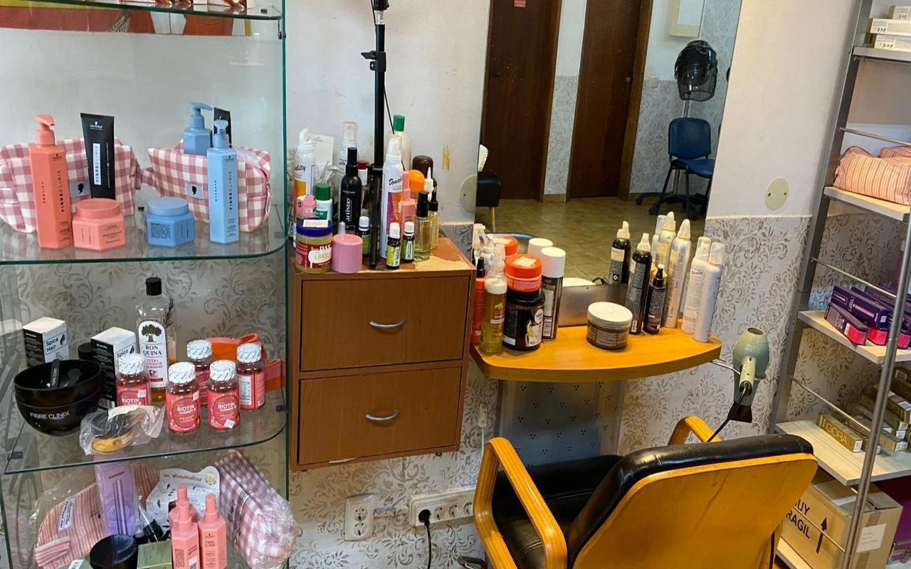 Le poste de travail d'un coiffeur avec un miroir mural et un plan de travail en bois encombré de produits coiffants, une vitrine de produits de soin capillaire et de compléments à côté, et un fauteuil de coiffure classique en bois et noir.