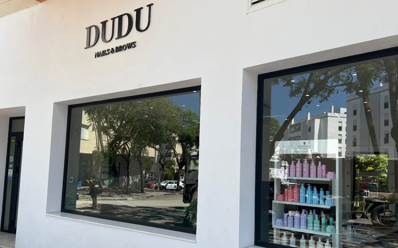 La devanture blanche de Dudu Nails and Brows à Estepona, avec son nom en lettres foncées au-dessus de larges vitrines présentant des rangées de produits capillaires.