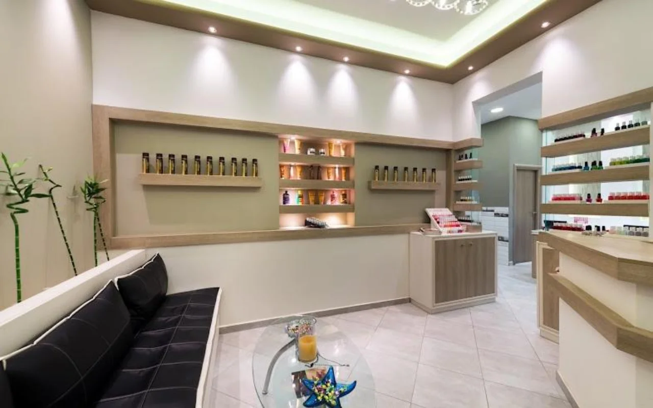 L'accueil aux tons crème de Fabulous Nails and More à Fira, Santorin, avec une banquette en cuir noir, une table en verre, des étagères rétroéclairées de produits de beauté et un éclairage doux au plafond.
