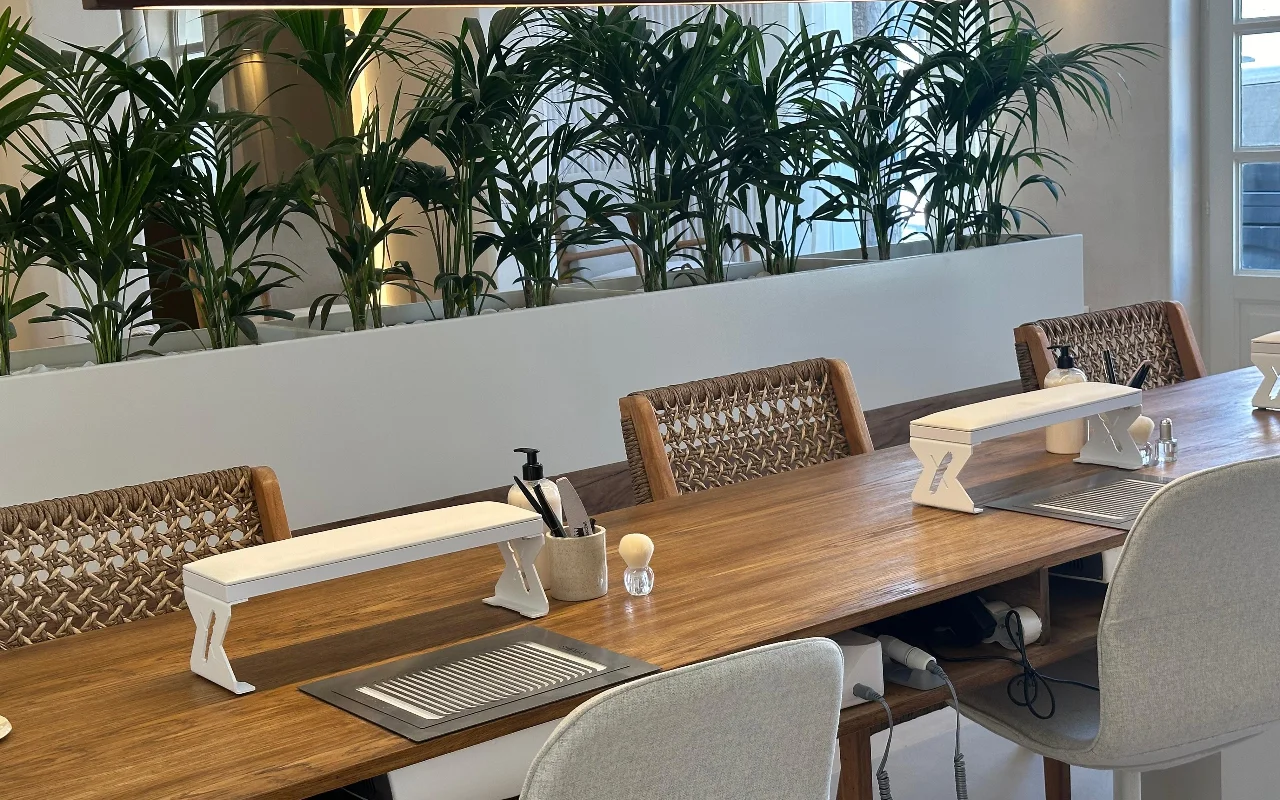 Une rangée de tables de manucure en bois chez Her Paros à Naoussa, chacune avec un repose-bras blanc et une chaise en rotin tressé, devant de grands palmiers verts et un mur miroir.