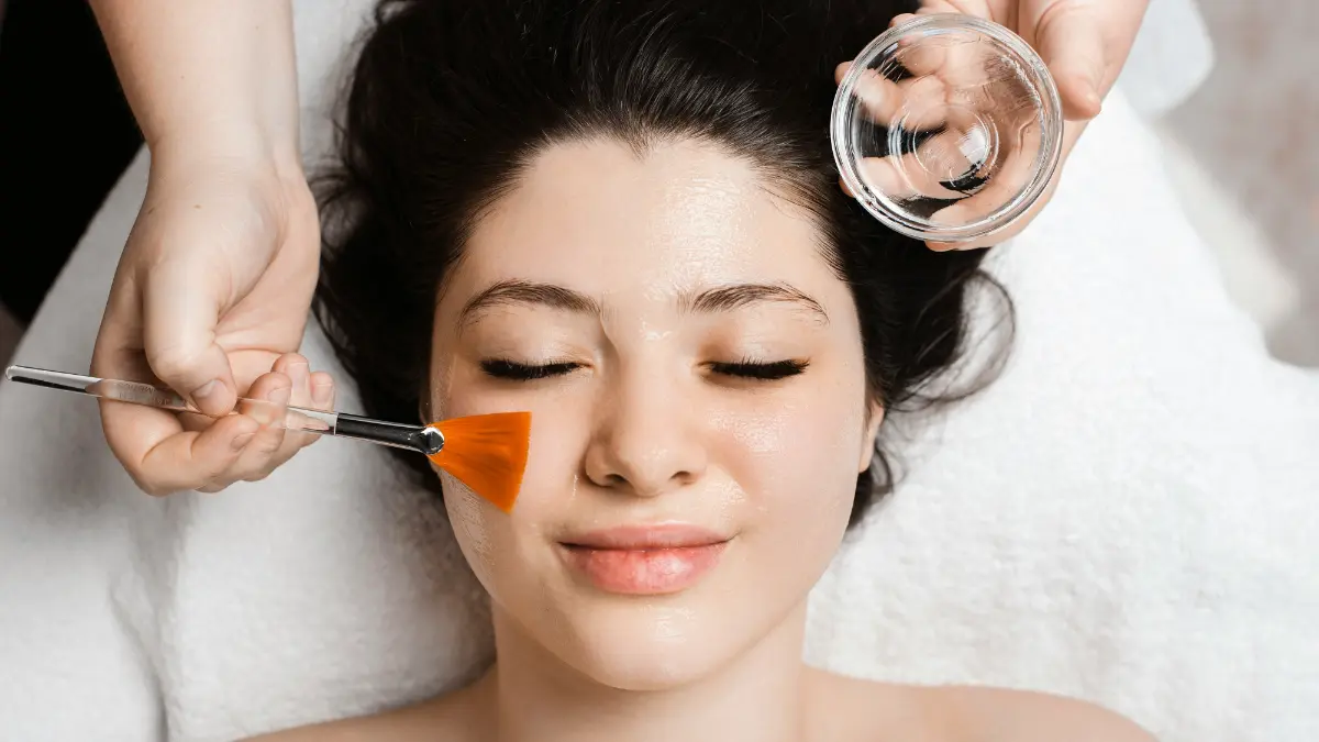 Vue de dessus du visage d'une cliente aux yeux fermés, pendant qu'un praticien applique au pinceau un masque apaisant