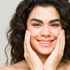 Une jeune femme à la peau nette et aux cheveux foncés ondulés sourit face à la caméra