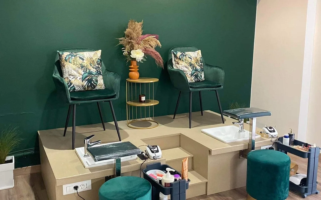 Un coin pédicure avec un mur vert profond, deux fauteuils tonneau en velours vert avec coussins à imprimé tropical surélevés sur une plateforme beige au-dessus de bassines pédicure, un guéridon doré tenant des herbes de pampa dans un vase orange, et des tabourets assortis en velours vert.