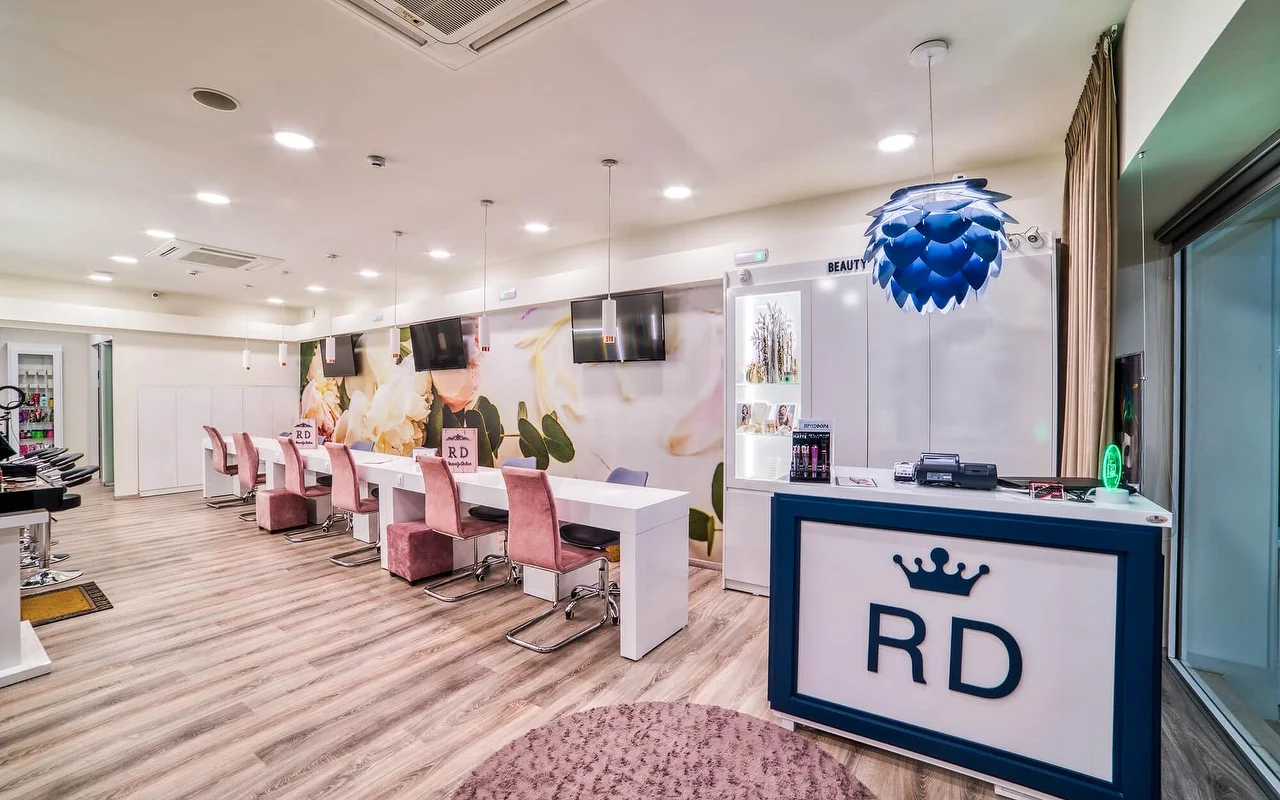 Un salon blanc et lumineux avec une longue rangée de tables de manucure et des chaises rose pâle, une douce fresque florale murale, un sol en bois clair, et un comptoir d'accueil marqué du logo couronne 'RD'.