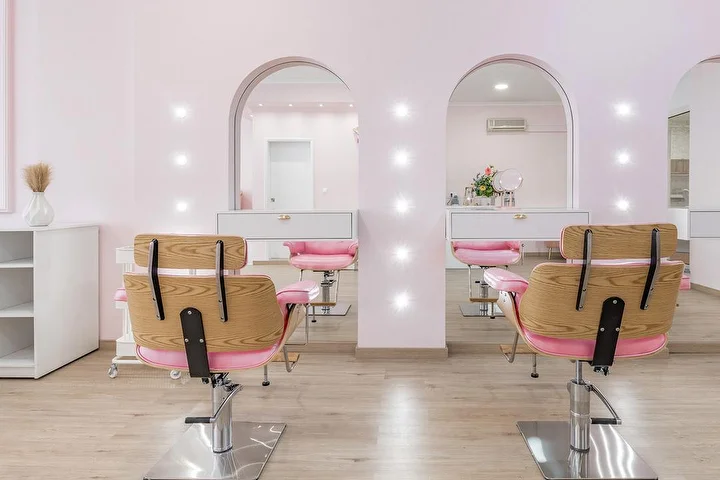 Salon de coiffure lumineux en rose pâle, avec des fauteuils de coiffage en bois et tissu rose face à deux niches en arche éclairées par des spots, meubles blancs et sol effet bois clair.