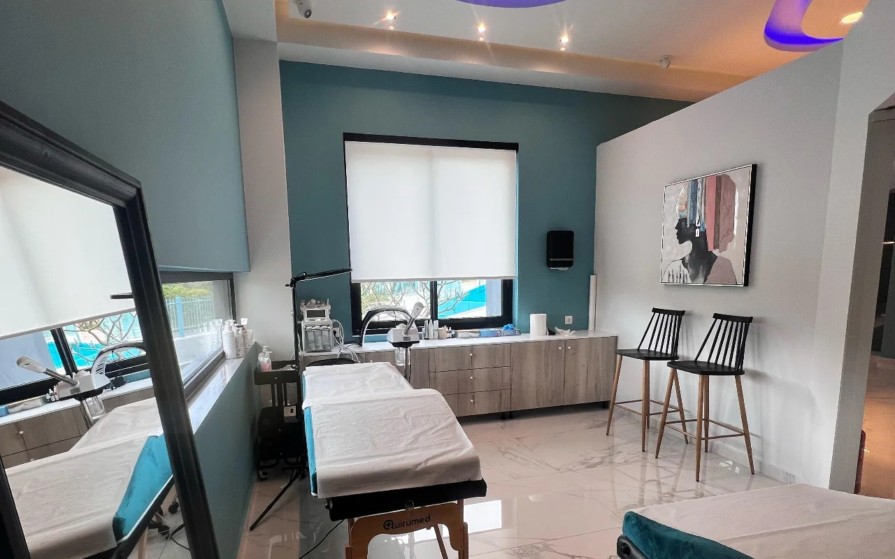 Une salle de soins visage avec des murs bleu canard, un lit de soins blanc, des appareils skincare, un grand miroir, deux tabourets noirs à lattes et un portrait encadré, éclairée par une fenêtre avec un store enrouleur blanc.