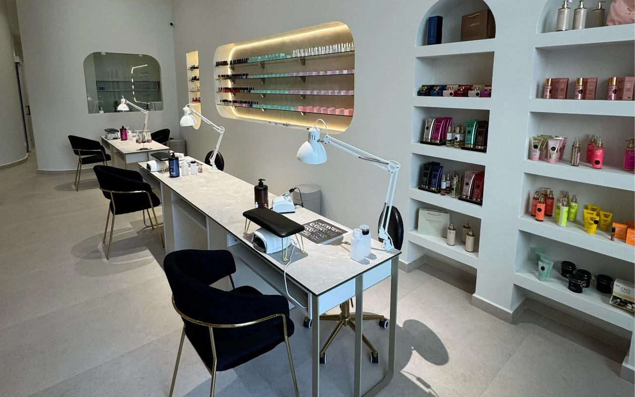 Un studio d'ongles blanc avec des tables de manucure au plateau de marbre et des lampes articulées blanches, des chaises en velours noir sur pieds dorés, et des niches murales en arc exposant des produits skincare et un présentoir rétroéclairé de vernis à ongles.