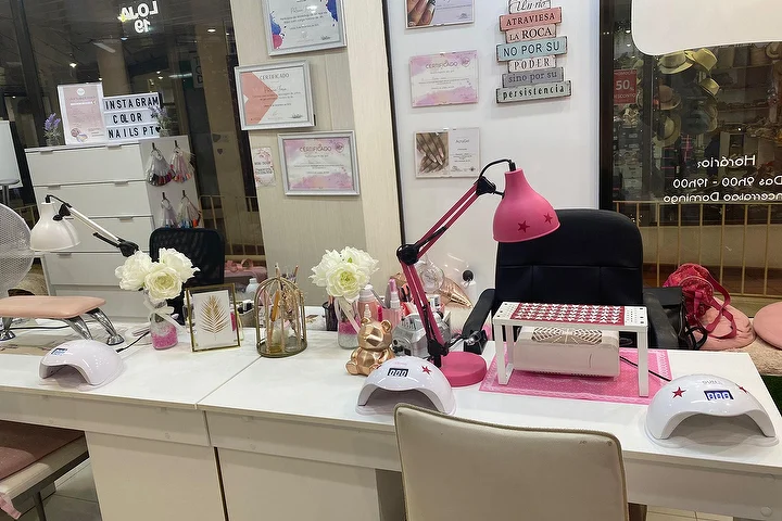 Poste de manucure d'un salon d'ongles avec des bureaux blancs, une lampe rose vif et des lampes LED, des roses blanches et des certificats encadrés au mur, avec le nom Color Nails PT visible sur la vitrine à côté des horaires d'ouverture.