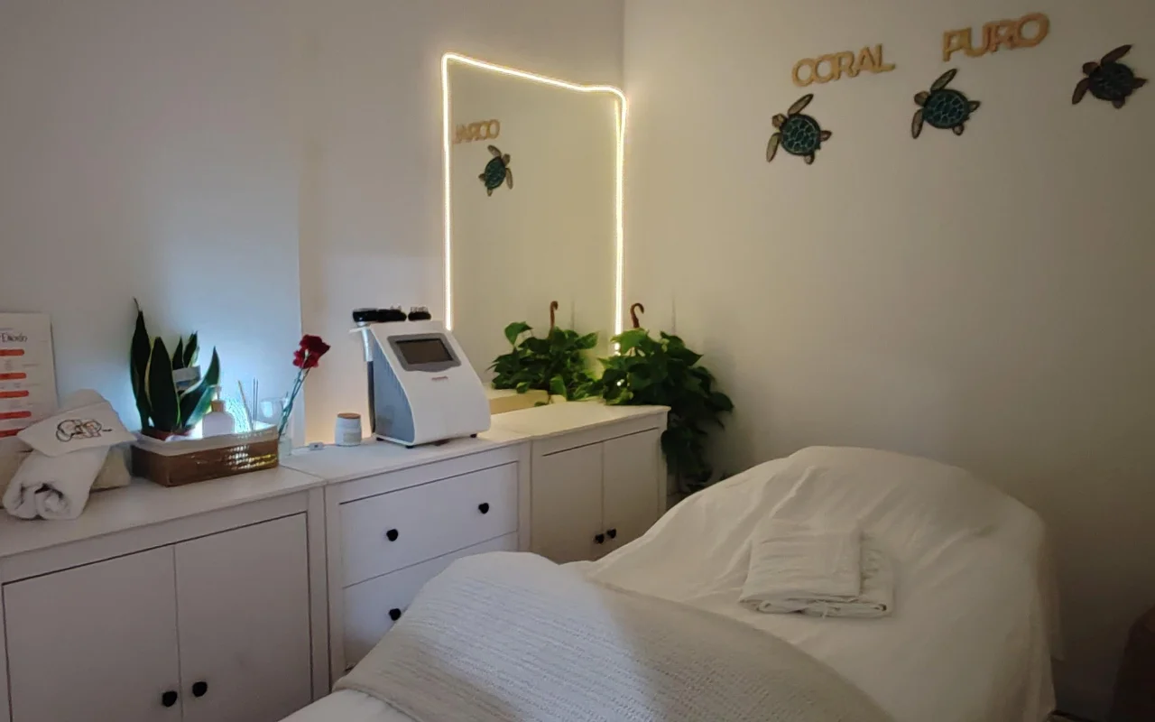 Une petite salle de soin calme avec des placards blancs et un lit habillé de serviettes blanches. Un miroir illuminé et une machine de soin trônent sur le comptoir parmi des plantes vertes, sous le lettrage doré « Coral Puro » et une décoration murale en forme de tortue.