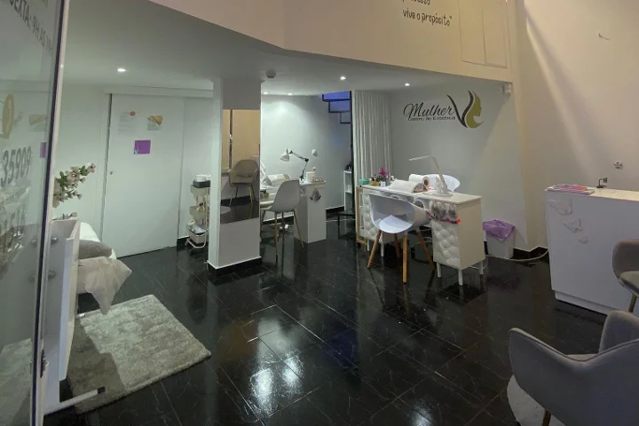Intérieur d'un centre de beauté : sol carrelé foncé et poli, poste de manucure blanc et espaces de soin doucement éclairés, logo Mulher au fond de la salle et une cabine de soin visage visible derrière une cloison en verre.