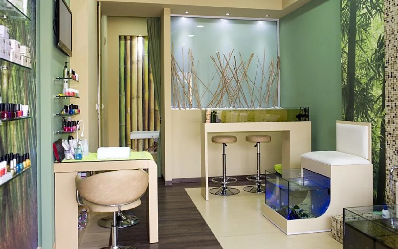 Un salon compact avec un décor mural en bambou et vert forêt, des étagères murales alignées de vernis à ongles, une table et une chaise de manucure crème, deux tabourets de bar, et un aquarium en verre éclairé de bleu pour la pédicure fish spa.