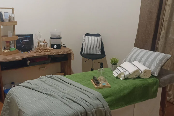 Cabine de massage chaleureuse avec une table drapée d'une serviette verte et de serviettes blanches roulées, un plateau d'huiles essentielles, des outils de massage en bois sur une petite table rustique, et un tabouret à côté d'un coin séparé par un rideau.