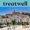 Vue ensoleillée sur Ibiza Ville, avec des bâtiments blanchis à la chaux qui grimpent jusqu'à la cathédrale et aux anciennes murailles de Dalt Vila sur la colline. Une promenade portuaire bordée d'arbres traverse le premier plan sous un ciel bleu limpide.