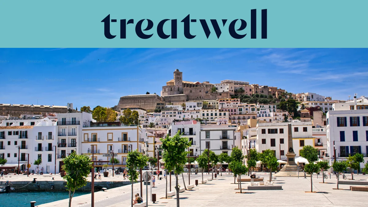 Vue ensoleillée sur Ibiza Ville, avec des bâtiments blanchis à la chaux qui grimpent jusqu'à la cathédrale et aux anciennes murailles de Dalt Vila sur la colline. Une promenade portuaire bordée d'arbres traverse le premier plan sous un ciel bleu limpide.