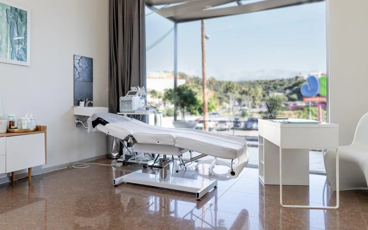 Une salle de soins blanche et épurée avec un lit esthétique inclinable, du matériel skincare sur un chariot, des œuvres encadrées et une grande fenêtre donnant sur des arbres, une route et des collines au loin.