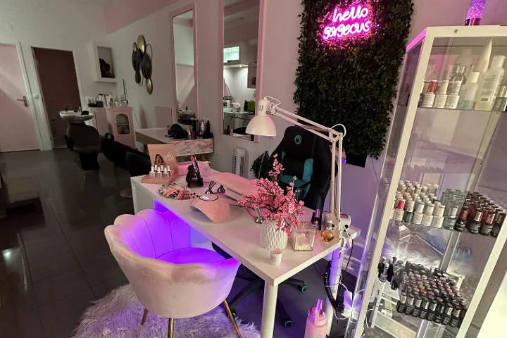 Poste beauté éclairé en rose, avec une enseigne néon 'hello gorgeous' sur un mur végétal, un fauteuil crème en forme de coquille, un bureau blanc avec pinceaux et fleurs de cerisier, et une vitrine remplie de vernis colorés.