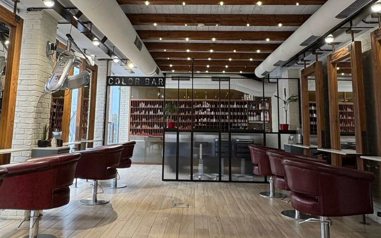 Un salon de coiffure avec des poutres en bois apparentes et de la brique blanchie à la chaux, des fauteuils de coiffage en cuir bordeaux, et une salle couleur aux parois vitrées bordée d'étagères de produits derrière une enseigne 'Color Bar'.