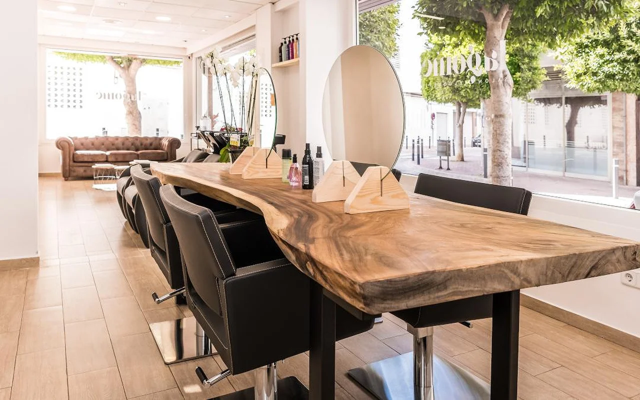 Un salon de coiffure lumineux construit autour d'un long comptoir en bois brut bordé de fauteuils de coiffure noirs. Un miroir rond et des présentoirs en bois trônent sur le comptoir, un canapé Chesterfield en cuir marron attend au fond, et de hautes fenêtres donnent sur une rue arborée.