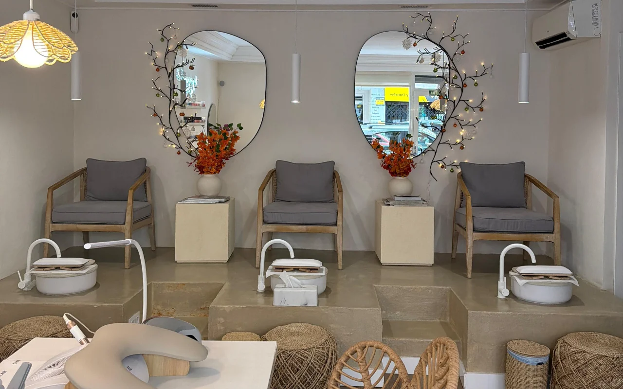 Un salon d'ongles moderne et lumineux avec deux fauteuils de pédicure sur une plateforme en béton surélevée, chacun équipé d'un bain de pieds blanc et d'un fauteuil rembourré gris. Deux miroirs aux formes organiques, ornés de branches lumineuses et de fleurs oranges, sont accrochés au-dessus, avec un poste de manucure au premier plan.