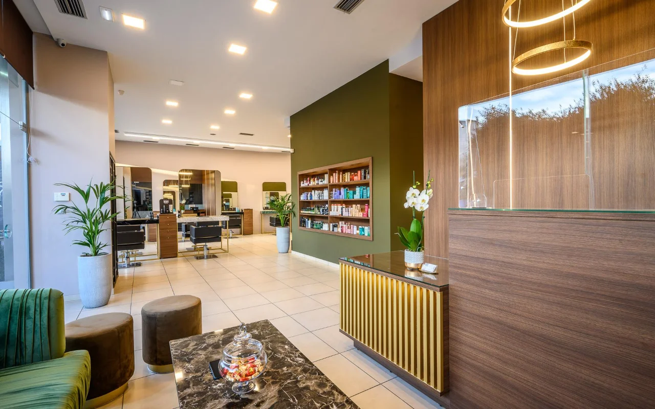 Un salon de coiffure haut de gamme avec des lambris de bois chaud, un mur d'appoint vert olive d'étagères produits, des postes de coiffage aux grands miroirs, un comptoir d'accueil à lattes dorées et des assises en velours vert sous des suspensions dorées circulaires.