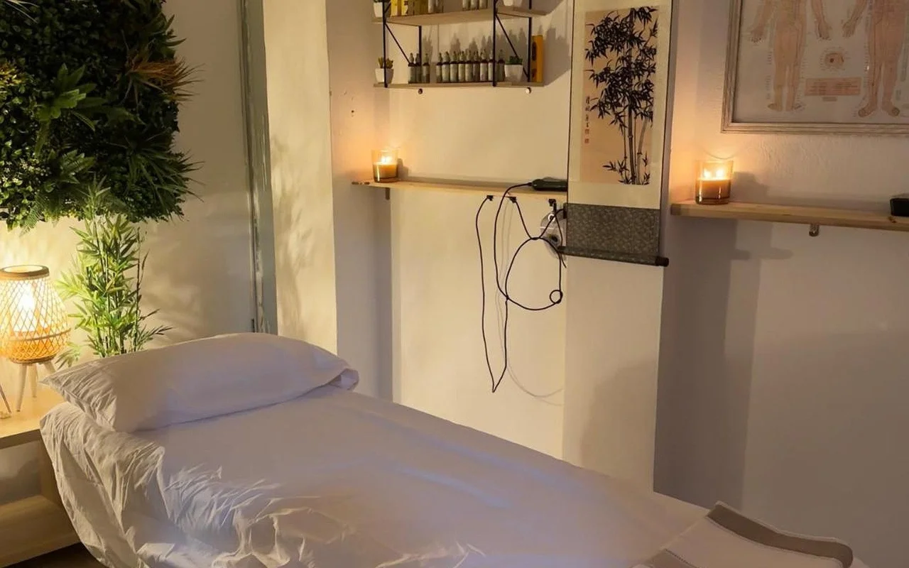 Une salle de soin faiblement éclairée avec une table de massage habillée de lin blanc. Des bougies allumées reposent sur des étagères murales en bois à côté d'un rouleau à l'encre en bambou et d'un schéma d'acupuncture encadré, avec une plante verte et une lampe en osier lumineuse dans le coin.