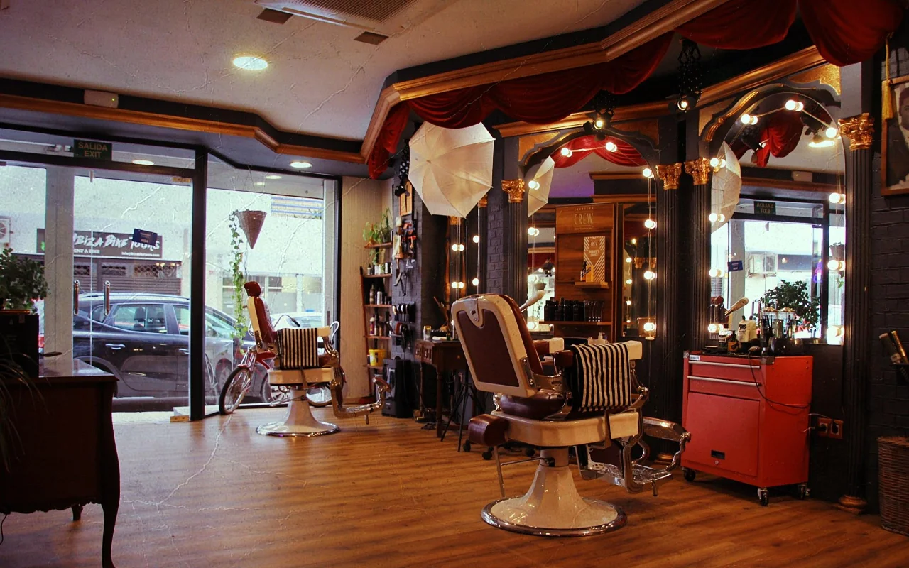 Un barbier au style vintage avec de classiques fauteuils inclinables en cuir sur bases chromées et des serviettes rayées. Des miroirs dorés encadrés d'ampoules rondes et des drapés de velours rouge tapissent le mur, un chariot à outils rouge se tient à côté, et des fenêtres côté rue éclairent le plancher en bois.