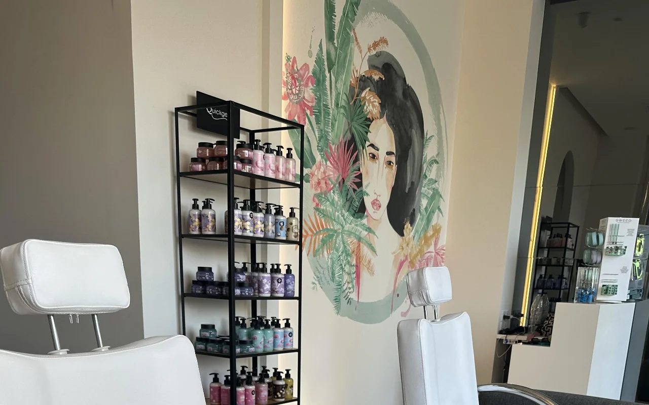 Un coin studio avec deux fauteuils de soins blancs inclinables, une étagère noire de produits, un miroir rétroéclairé, et une fresque florale murale représentant un visage de femme entouré d'un feuillage tropical.