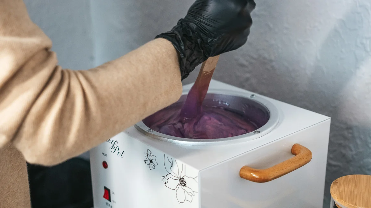 La main d’un praticien, gantée de noir, mélange un pot de cire chaude fondue dans un institut de beauté