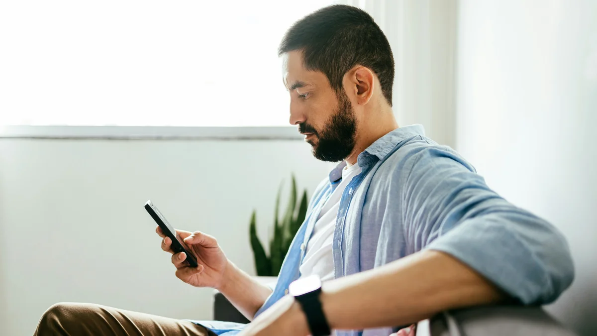 Vue de côté d’un homme assis sur son canapé, un bras posé sur le dossier, qui réserve un soin sur son téléphone de l’autre main