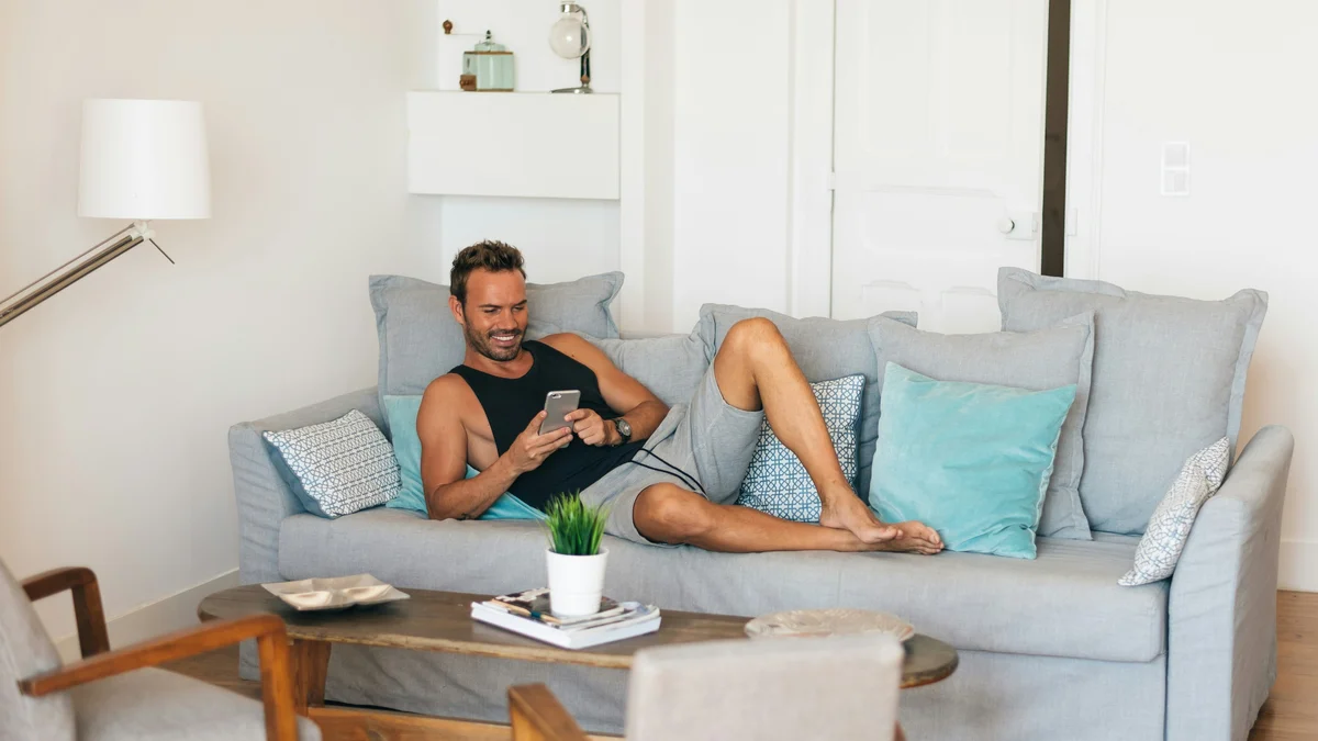 Un homme se détend sur son canapé, les pieds surélevés, en souriant tout en regardant son téléphone ; il porte un débardeur noir et un short gris ample et doux