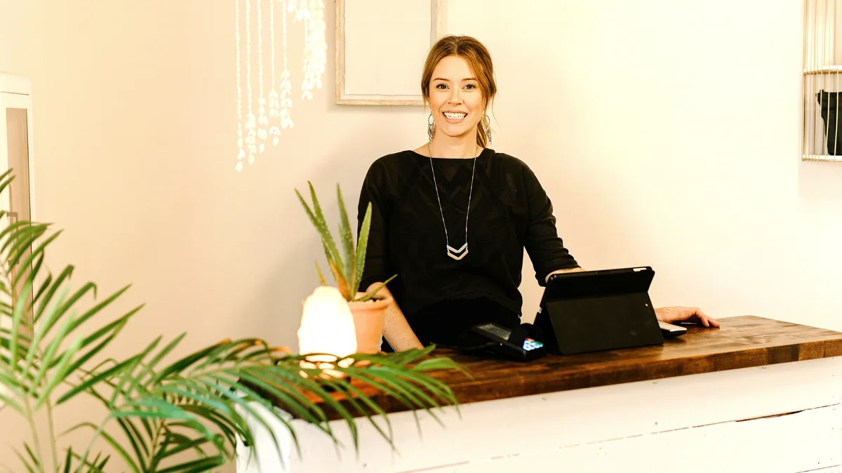 Une femme souriante se tient derrière le comptoir d’accueil d’un institut de beauté